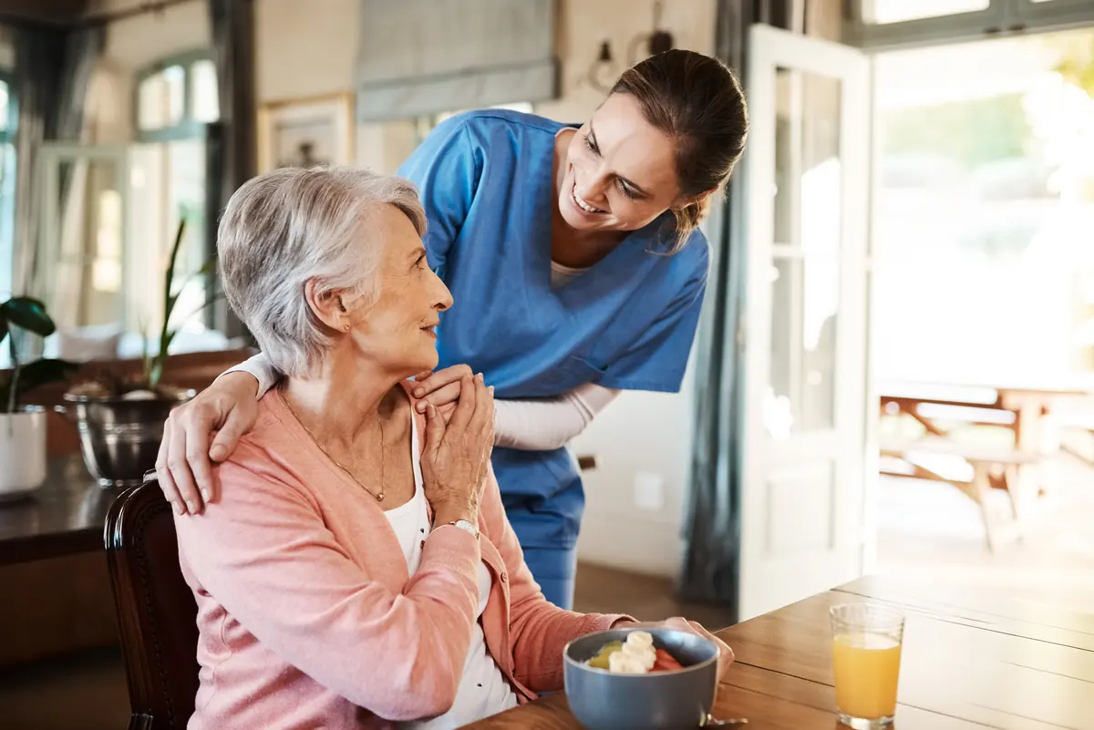 A nurse in blue scrubs leaning in to talk with an older woman seated at a table at home